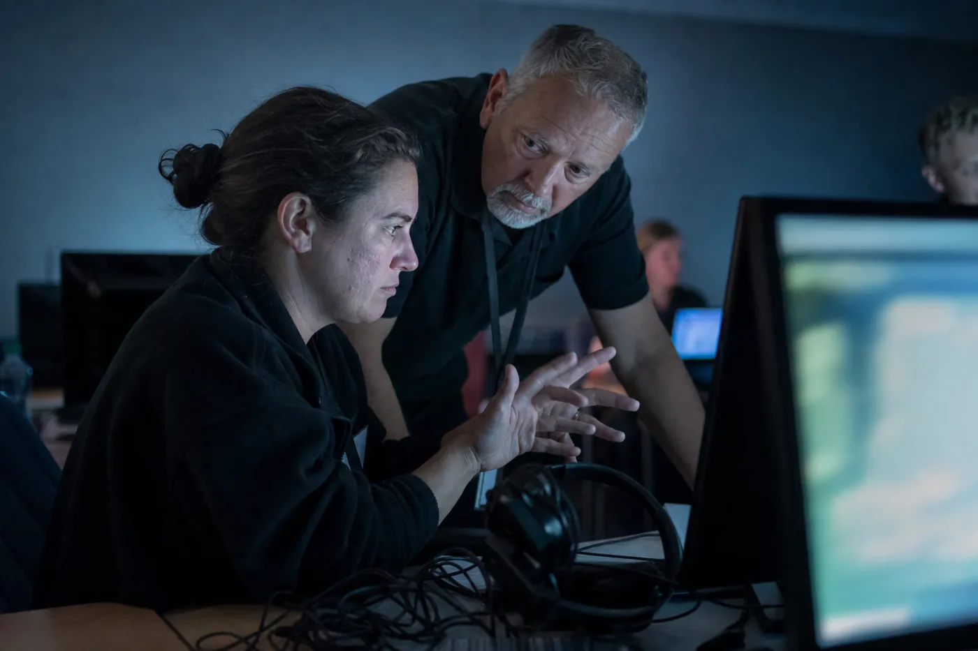 A trainee practicing a high-pressure decision at a workstation while an experienced colleague leans in to coach her through it