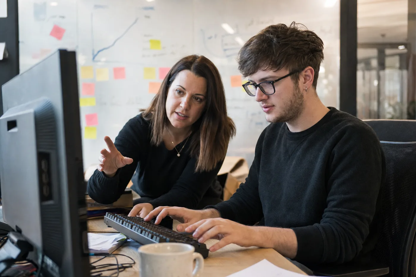 A manager coaching a colleague at his desk with a hand-drawn rising curve on a glass wall behind them