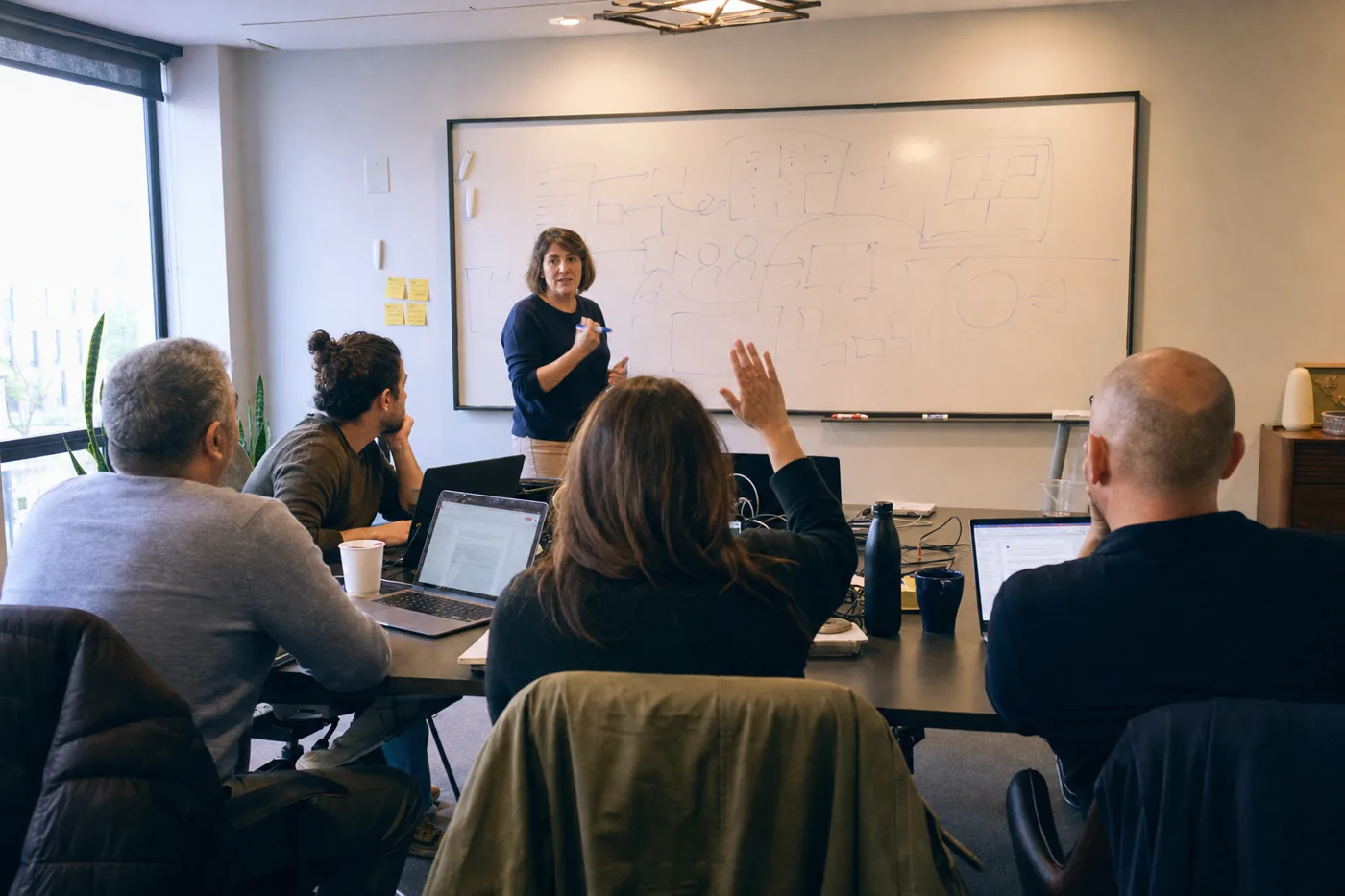 A facilitator leading a small employee training session at a whiteboard while a participant raises her hand