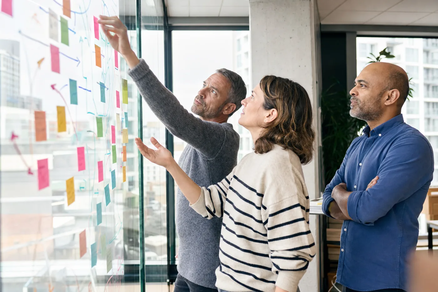 Coworkers mapping moments of need on a glass wall with colored sticky notes in bright overcast daylight, planning where learning should meet the work